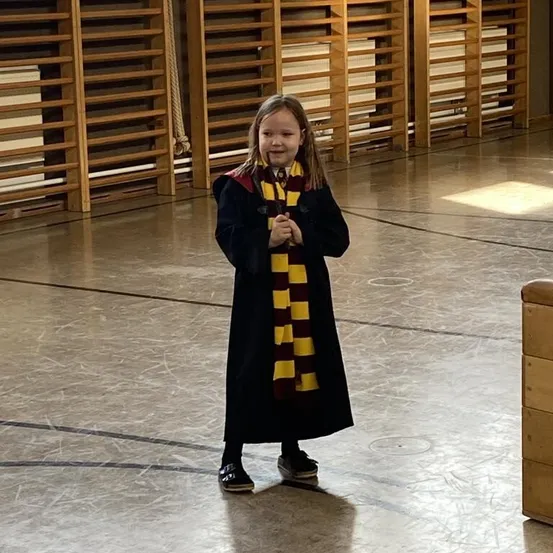 Ein junges Mädchen in einem Gryffindor-Umhang und -Schal steht in einem Raum mit Holzwänden. Sie schaut lächelnd nach oben.