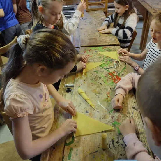 Mehrere junge Mädchen arbeiten in einem Klassenzimmer an einem Bastelprojekt. Sie sitzen an Tischen, die mit Farbe bedeckt sind, wobei einige von ihnen Scheren halten und andere gelbes Papier in der Hand haben.