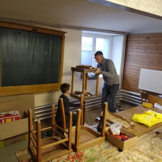 Ein Mann und ein Kind bauen Holzstühle in einem Raum zusammen. An der Wand steht eine Tafel und ein Heizkörper. Ein Tisch mit einer gelben Tasche steht daneben, und einige Kisten liegen auf dem Boden.