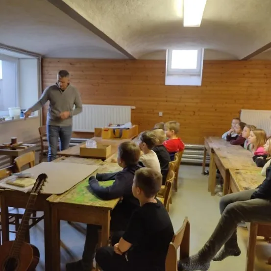 Ein Klassenzimmer mit Holzwänden und Tischen, an denen Kinder sitzen. Ein Mann steht vor der Klasse. Eine Gitarre ist links und ein Heizkörper ist an der Wand.