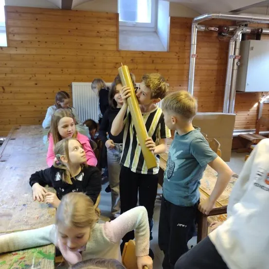 Eine Gruppe von Kindern ist um einen Tisch mit einem gelben Rohr in einem Klassenzimmer versammelt. Einige schauen darauf, während andere sitzen und beobachten. Der Raum hat Holzwände und einen weißen Heizkörper.