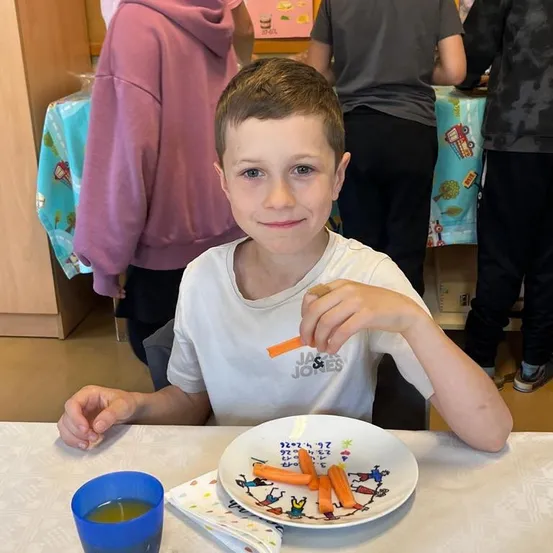 Ein Junge in einem weißen T-Shirt sitzt an einem Tisch und isst Karottensticks. Ein Teller mit Karotten und ein blauer Becher sind auf dem Tisch. Andere Leute stehen hinter ihm.