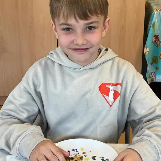 Ein junger Junge mit einem Lächeln sitzt an einem Tisch und hält einen Teller mit einem Keks. Er trägt eine graue Kapuzenjacke mit einem roten Superman-Logo. Der Hintergrund besteht aus einer Holzwand und einer bunten Tischdekoration.
