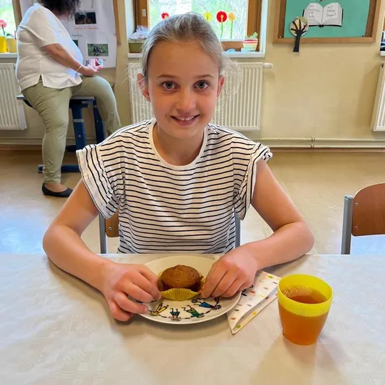 Ein junges Mädchen sitzt am Tisch und isst einen Muffin mit einem Getränk. Eine erwachsene Frau sitzt im Hintergrund.