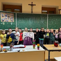 Ein Klassenfoto zeigt Kinder, die vor Tafeln mit der Aufschrift Erste Kommunion posieren und einem Kreuz über einer Tür.