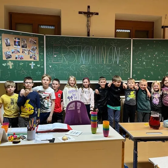 Ein Klassenfoto zeigt Kinder, die vor Tafeln mit der Aufschrift Erste Kommunion posieren und einem Kreuz über einer Tür.