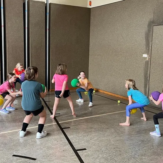 Mehrere junge Mädchen spielen eine Partie Dodgeball in einer Turnhalle. Sie halten farbige Bälle und tragen Sportkleidung. Der Raum hat einen Betonboden und Wände.