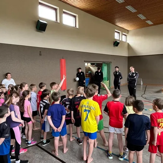 Eine Gruppe von Kindern und Erwachsenen in einer Turnhalle, ein Kind zeigt auf einen von einem Erwachsenen gehaltenen Fußball. Einige Erwachsene stehen und beobachten.