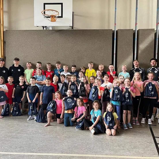 Eine Gruppe von Kindern und Erwachsenen steht zusammen in einer Sporthalle, lächelt und hält Taschen. Hinter ihnen ist ein Basketballkorb an der Wand montiert.