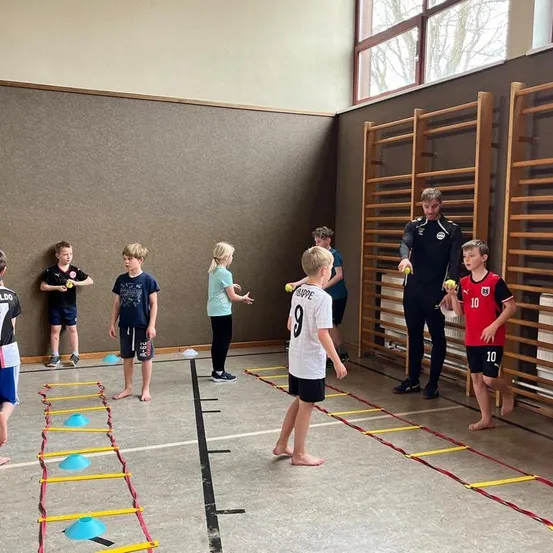 Eine Gruppe von Kindern in einem Fitnessstudio, die an einer Trainingseinheit mit einem Trainer teilnehmen. Sie halten Tennisbälle in den Händen und stehen vor Hüpfleitern.