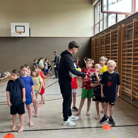 Eine Gruppe von Kindern steht in einer Turnhalle mit einem Mann in einer schwarzen Kapuzenjacke, der einen Fußball übergibt. Die Kinder sind barfuß.