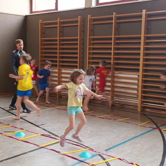 Kinder in einer Turnhalle spielen ein Spiel mit einem Trainer und springen über Kegel auf einem Teppich. Ein Mädchen führt, trägt ein gelbes Shirt und Shorts.
