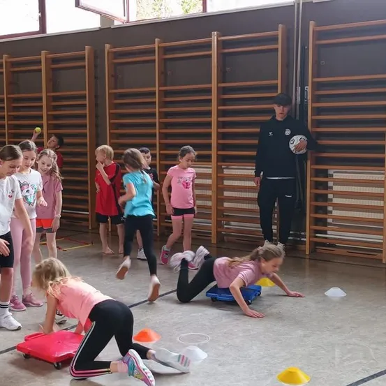 Mehrere Kinder spielen mit kleinen roten und blauen Wagen auf dem Boden. Einige Kinder beugen sich hinunter, während andere hinter hölzernen Stangen stehen. Ein junger Mann mit einem Ball steht hinter ihnen.