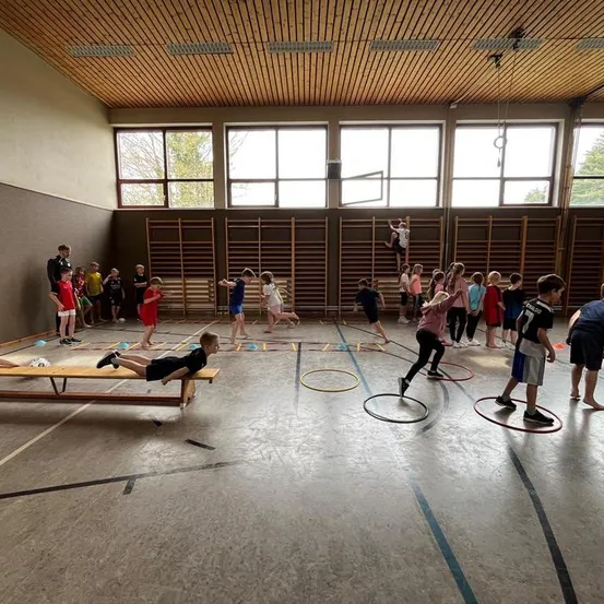 Kinder betreiben in einer Turnhalle körperliche Aktivitäten, einige am Boden, andere auf einer Bank, und Hula-Hoops verstreut. Einige klettern auf Holzstangen.