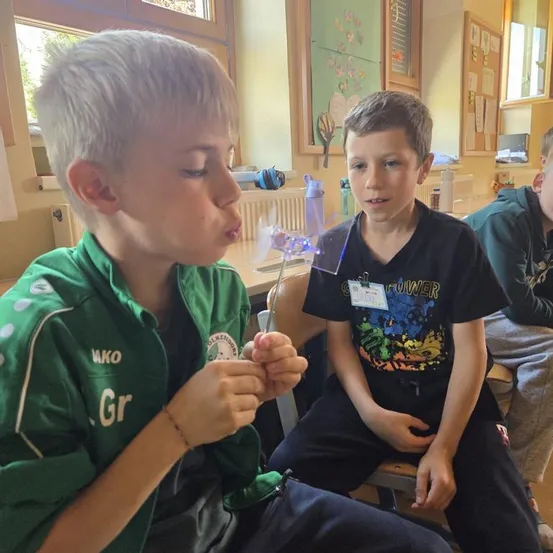 Zwei Jungen sitzen in einem Klassenzimmer, einer bläst auf ein Lichtspielzeug. Der Junge in Grün trägt ein grünes Trikot mit weißen Buchstaben. Der Junge in Schwarz hat ein bedrucktes Shirt und einen Namensschild. Ein dritter Junge sitzt hinter ihnen.