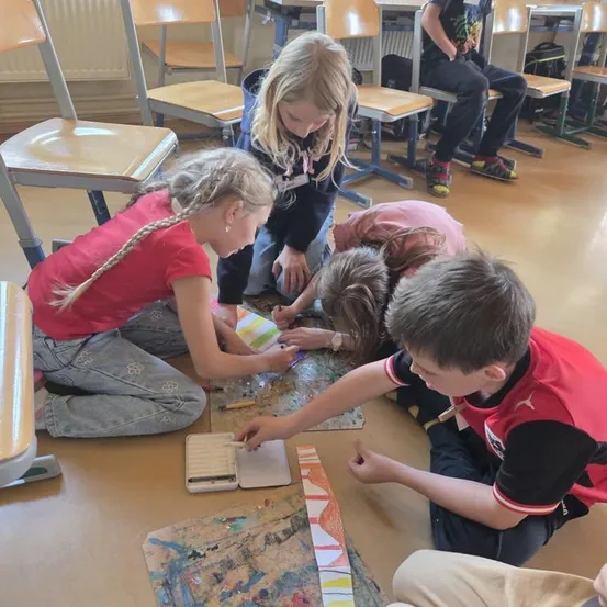 Kinder sitzen auf dem Boden und arbeiten an einem Kunstprojekt. Sie verwenden bunte Marker und Papier. Andere Kinder sitzen auf Stühlen im Hintergrund.