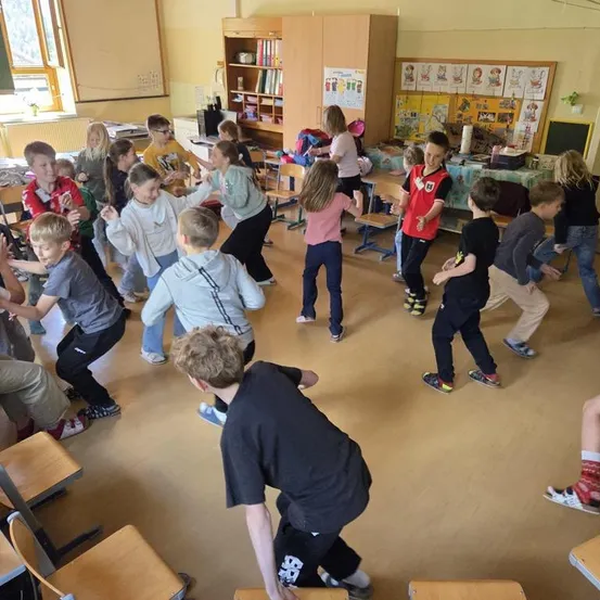 Ein Klassenzimmer voller Kinder, die sich mit verschiedenen Aktivitäten beschäftigen. Einige tanzen, andere sitzen. Im Hintergrund befinden sich Regale mit Büchern und eine Pinnwand.