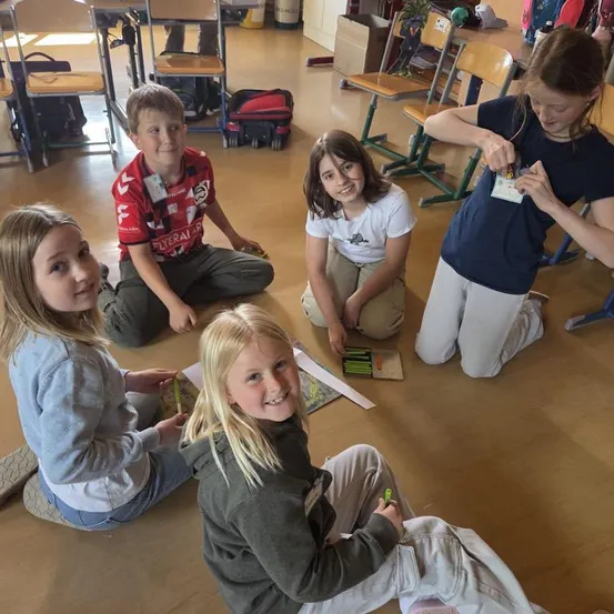 Fünf Kinder sitzen lächelnd auf dem Boden und arbeiten an einem Kunstprojekt. Ein Kind hält einen Abzeichen. Sie haben Bücher und einen grünen Stift in der Nähe. Der Raum hat Holzböden und Stühle.