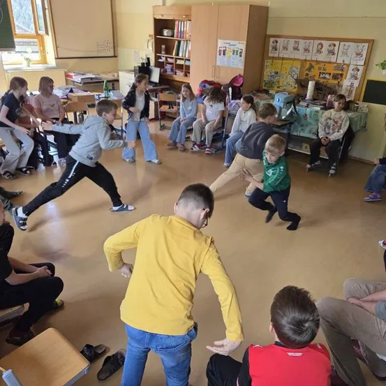 Eine Gruppe von Kindern ist in einem Klassenzimmer und macht eine Übung. Einige Kinder stehen und machen Dehnübungen, während andere auf Stühlen sitzen. Es gibt einen Tisch mit Büchern und anderen Gegenständen.
