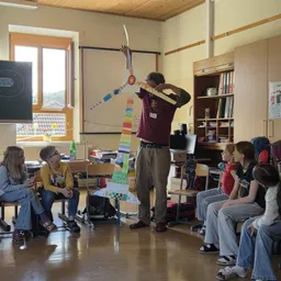 Ein Mann hält ein großes Windradmodell in einem Klassenzimmer. Kinder sitzen um ihn herum und lauschen aufmerksam.
