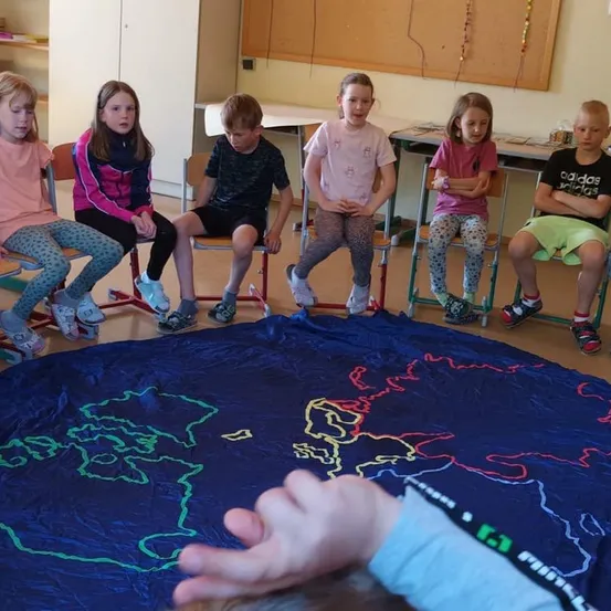 Eine Gruppe von Kindern sitzt in einem Klassenzimmer mit einer großen Weltkarte auf dem Boden. Einige hören aufmerksam zu, während andere desinteressiert erscheinen.