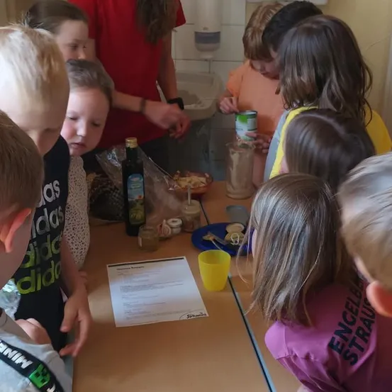 Kinder in einem Klassenzimmer sind um einen Tisch versammelt. Sie schauen sich ein Rezept an und haben verschiedene Gegenstände wie Flaschen, Becher und Teller auf dem Tisch.
