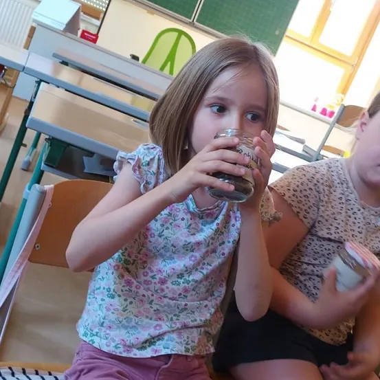 Zwei junge Mädchen sitzen an einem Schreibtisch in einem Klassenzimmer. Eines der Mädchen trinkt aus einem Glas, während das andere ein Glas mit einem roten und weißen Deckel hält.