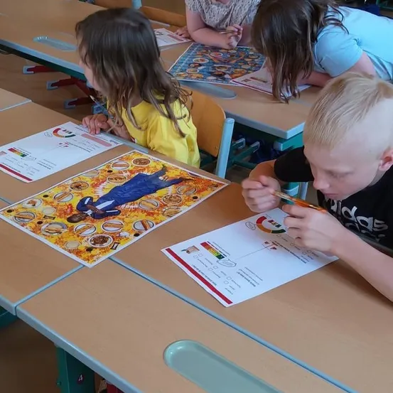 Schüler in einem Klassenzimmer arbeiten an Kunstprojekten. Ein Junge färbt ein großes Papier ein und ein Mädchen schaut auf ihr Werk. Andere Schüler sitzen hinter ihnen an Tischen.