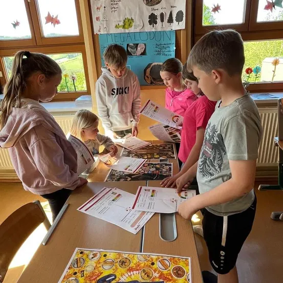 Eine Gruppe von Kindern, Jungen und Mädchen, steht um einen Tisch in einem Klassenzimmer und schaut sich Papiere und Bilder an. Ein Junge trägt eine Kapuzenjacke mit dem Wort 'KRANK!' darauf.