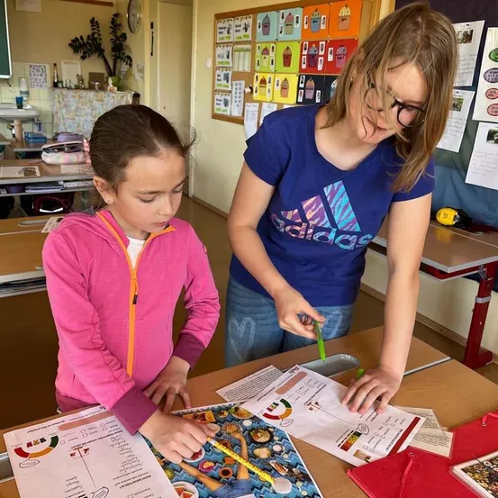 Zwei Mädchen in einem Klassenzimmer sind in einer Aktivität engagiert, eine hält einen Bleistift und die andere hält ein Papier mit einem Diagramm darauf. Der Tisch ist mit Papieren, einer roten Mappe und einem grünen Bleistift übersät. Im Hintergrund befinden sich Wände mit Postern und einem Regal mit Gegenständen.