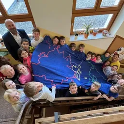 Eine Gruppe von Kindern und Erwachsenen versammelt sich um einen blauen Stoff mit einer Weltkarte darauf. Sie lächeln und posieren für ein Foto. Der Raum hat gelbe Wände und Holzgeländer.