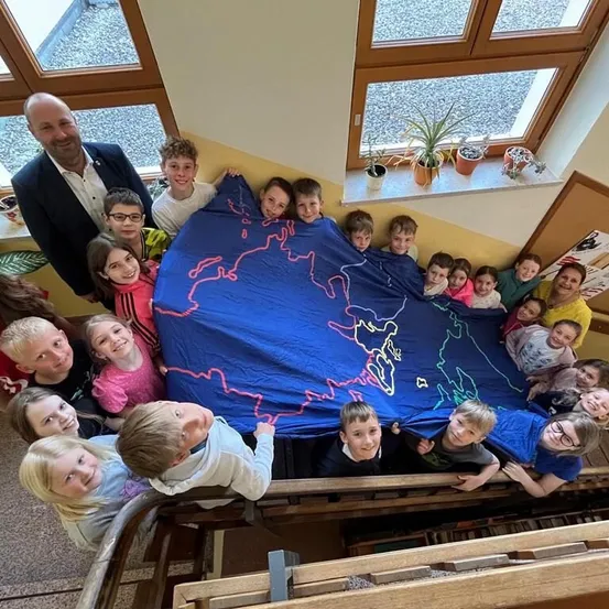 Eine Gruppe von Kindern und Erwachsenen versammelt sich um einen blauen Stoff mit einer Weltkarte darauf. Sie lächeln und posieren für ein Foto. Der Raum hat gelbe Wände und Holzgeländer.