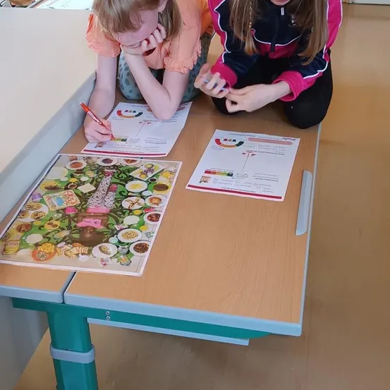 Zwei junge Mädchen arbeiten an einem Tisch an einer Ernährungsaktivität, eine schreibt, während die andere einen bunten Ernährungsplan betrachtet.