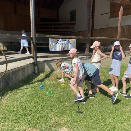 Eine Gruppe von Kindern spielt mit bunten Ringen auf einem Rasenfeld, mit einer hölzernen Bühne und einem Banner im Hintergrund.