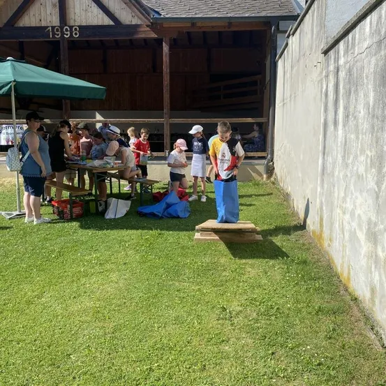 Eine Gruppe von Menschen, einschließlich Kindern, versammelt sich auf einer Rasenfläche. Ein Junge steht auf einer Holzplattform, während andere an einem Tisch unter einem Regenschirm sitzen. Ein Gebäude mit der Nummer 1998 ist im Hintergrund zu sehen.