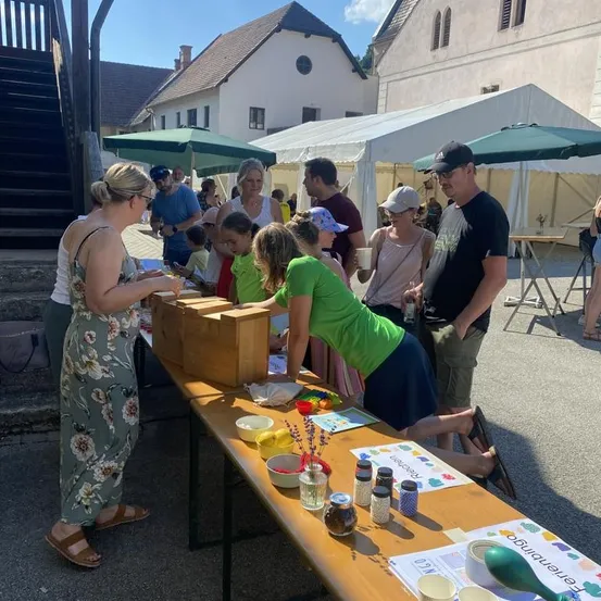 Eine Gruppe von Menschen versammelt sich um einen Tisch mit verschiedenen Gegenständen, möglicherweise bei einem Festival. Eine Frau in einem grünen Shirt beugt sich über den Tisch.