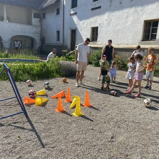 Eine Gruppe von Kindern und Erwachsenen spielt ein Spiel mit Bällen und Kegeln auf einem Kiesplatz vor einem Gebäude.