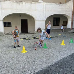 Kinder spielen Tennis auf einem Außenhof, eine Frau und ein Mädchen beobachten. Bunte Kegel und eine Leiter sind auf dem Boden verstreut.