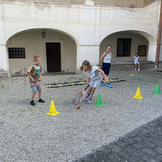 Kinder spielen Tennis auf einem Außenhof, eine Frau und ein Mädchen beobachten. Bunte Kegel und eine Leiter sind auf dem Boden verstreut.