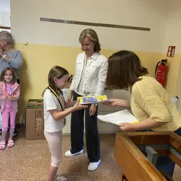 Ein junges Mädchen erhält von einer Frau in einer weißen Jacke ein Buch, während eine andere Frau ein Papier hält. Ein Mädchen und eine ältere Frau stehen in der Nähe.