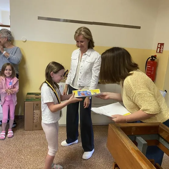 Ein junges Mädchen erhält von einer Frau in einer weißen Jacke ein Buch, während eine andere Frau ein Papier hält. Ein Mädchen und eine ältere Frau stehen in der Nähe.
