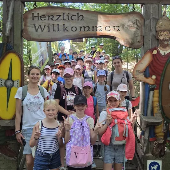 Eine Gruppe von Kindern und Erwachsenen posiert für ein Foto vor einem Holzschild mit der Aufschrift Herzlich Willkommen. Sie tragen Hüte und haben Rucksäcke dabei.