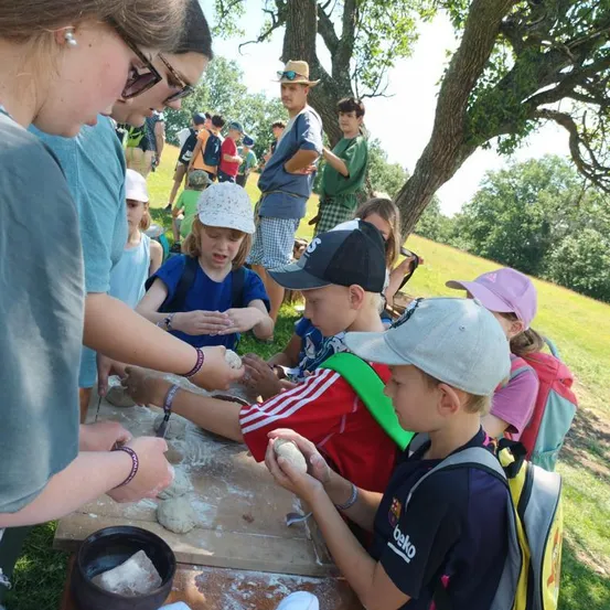 Eine Gruppe von Kindern und Erwachsenen versammelt sich in einem Park und ist mit einer Töpferaktivität beschäftigt. Sie sitzen um einen Tisch herum, einige von ihnen tragen Hüte und Rucksäcke. Der Tisch ist mit Ton und Werkzeugen übersät, und im Hintergrund befinden sich Bäume und ein Grasfeld.