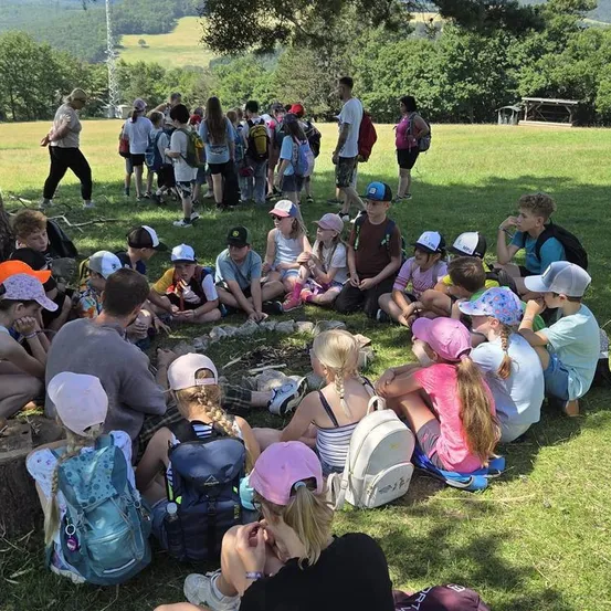 Eine Gruppe von Kindern und Erwachsenen sitzt im Kreis auf dem Gras, einige tragen Mützen und Rucksäcke. Sie scheinen in eine Diskussion vertieft zu sein. Im Hintergrund ist ein Grasfeld mit Bäumen und einem Turm zu sehen.