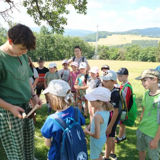 Eine Gruppe von Kindern in Hüten und Rucksäcken versammelt sich um einen Mann, der ein Stück Holz in einem Grasfeld hält, mit einer malerischen Aussicht auf Berge und Bäume im Hintergrund.