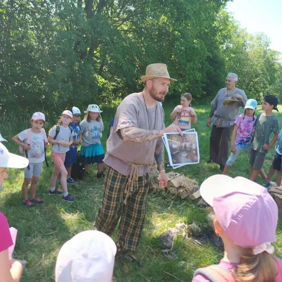 Ein Mann in einem Strohhut und karierten Hosen zeigt einem Kindergruppen ein Bild auf einer Rasenfläche mit Bäumen im Hintergrund.