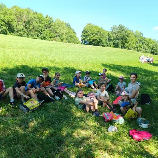 Eine Gruppe von Kindern und Erwachsenen sitzt auf einem Grasfeld unter einem Baum, einige tragen Hüte und Turnschuhe, und sie sind von Rucksäcken und anderen Gegenständen umgeben.