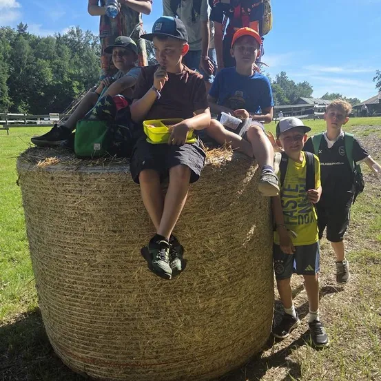 Eine Gruppe von Jungen sitzt auf einem großen Heuballen in einem Feld, mit einigen, die dahinter stehen. Sie tragen Freizeitkleidung und einige halten Snacks. Der Hintergrund zeigt Bäume und einen klaren blauen Himmel.