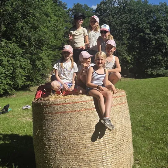 Eine Gruppe junger Mädchen in Hüten sitzt auf einem großen Heuballen in einem Grasfeld mit Bäumen im Hintergrund.