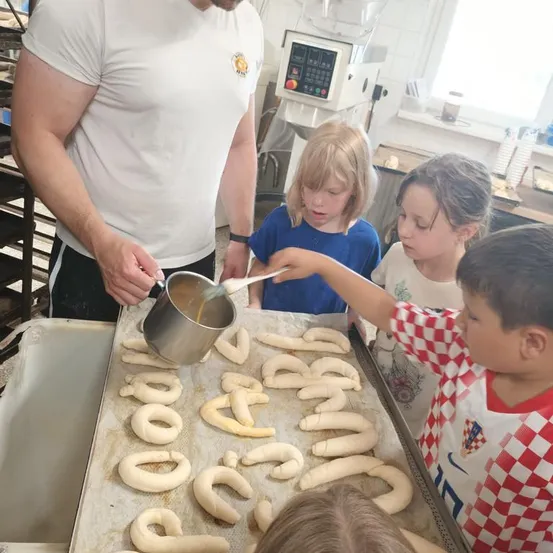 Ein Mann in einem weißen T-Shirt gießt Sirup auf Brezeln, während Kinder zusehen und helfen. Die Kinder stehen um eine Tablett mit Brezeln, die Buchstaben formen.